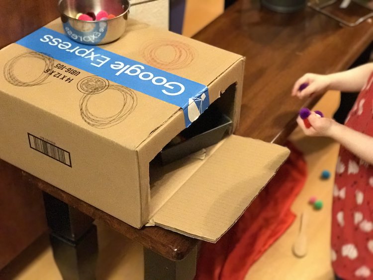 Cardboard box with a stovetop colored on top holding a pot full of pompoms. The front is opened to show a loaf pan about to come out or go in.