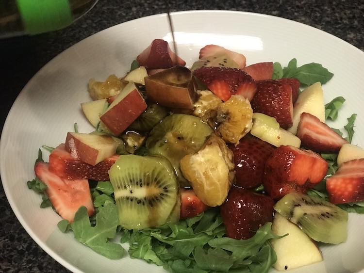 A white bowl filled with lettuce, diced fruit, with a drizzle of dark dressing being poured overtop.