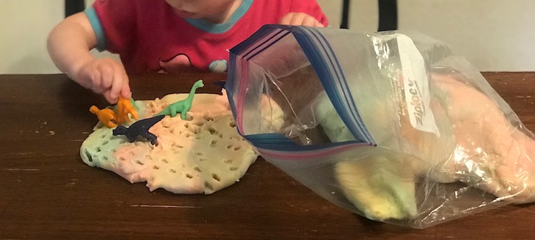 Image shows a circle of playdough on the table with a bag full beside it. Zoey is currently placing a dinosaur into the playdough. Three other dinosaurs are already there with tons of footprints around. 