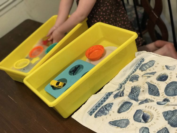 Ada washing her new sand toys along with a couple dinosaurs and cars. The wash bin is in the back with the plain water bin in front of it. Closest to the front, and cut off, is a kitchen towel.