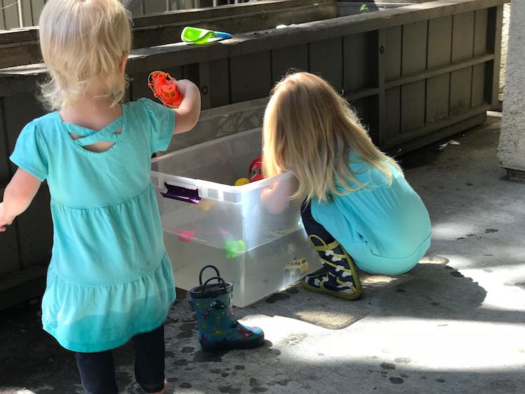Showing the back of Ada and Zoey. Ada is crouched in front of the plastic bin of water while Zoey brings a boat over to the bin.