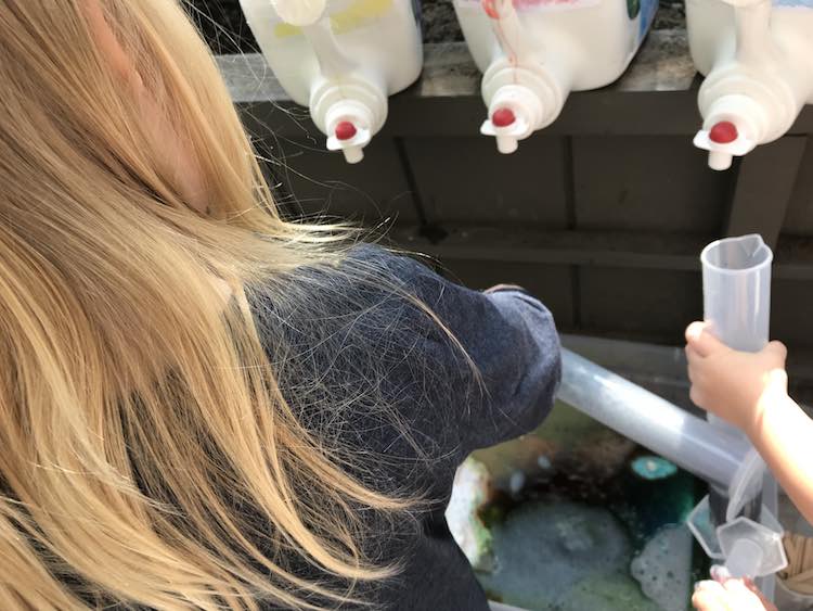 The back of Ada's hair is seen as she pours her graduated cylinder out into the bin below. Zoey is off photo but you can see her hand holding her own graduated cylinder as it approaches the empty laundry jugs filled with dyed vinegar.