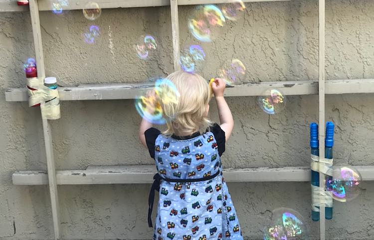 Back of Zoey is show pulling the want out of the bubble mix. Between her and the camera are several bubbles floating in the air.