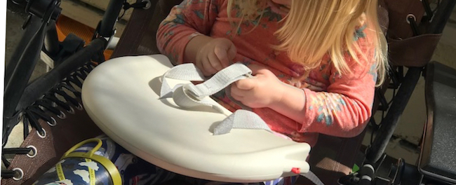 Ada sits in the chair focused on the three point buckle system on the highchair's back rest.