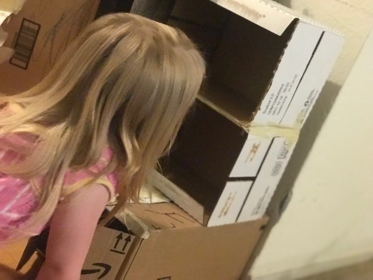 Ada is kneeling down looking into the shelving. The Soylent boxes are now stacked on each other and balanced near the back of the cupboard box.
