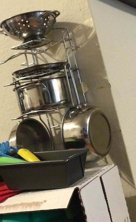 Closeup of the stand again but this time it’s covered in pots, lid, and strainer. In front of the stand but still on the shelves sits a loaf pan with some plastic play utensils.