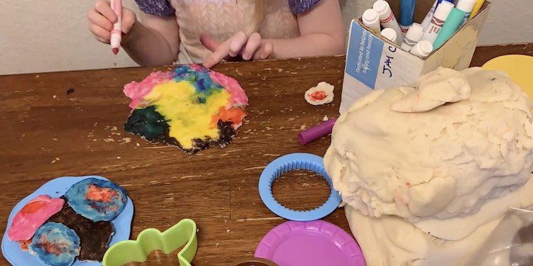 To the left is a plate of colorful playdough cookies. To the right is a giant pile of white playdough. Behind them is a closeup of Ada coloring a flat piece of playdough with washable markers token from the cardboard cup behind the playdough.