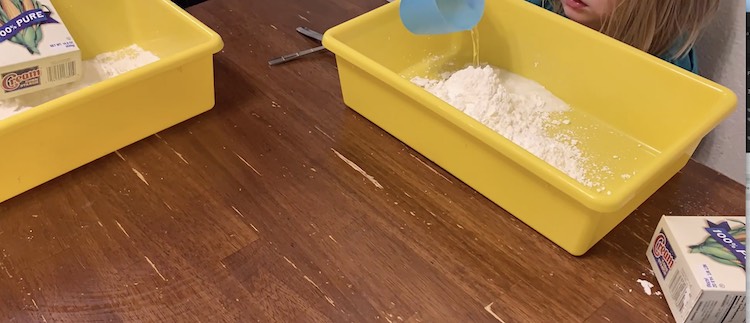 Zoey is still emptying her box of cornstarch into her sensory bin while Ada already did and is now pouring her cup of water into her bin.