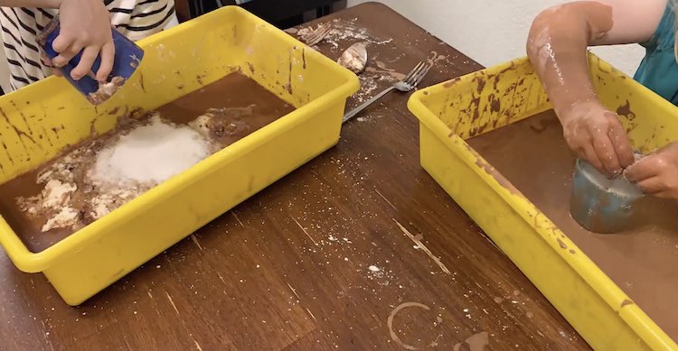 Zoey dumping her salt into her muddy oobleck while Ada adds the oobleck to her cup full of salt.