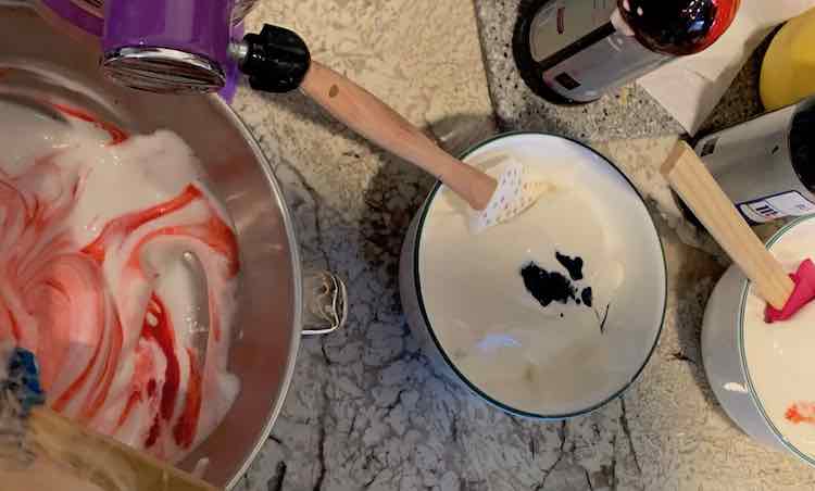 Partially mixed red angel food cake in the stand mixer. Two other bowls of angel food cake in the background with blue and yellow food dye dots in it. 