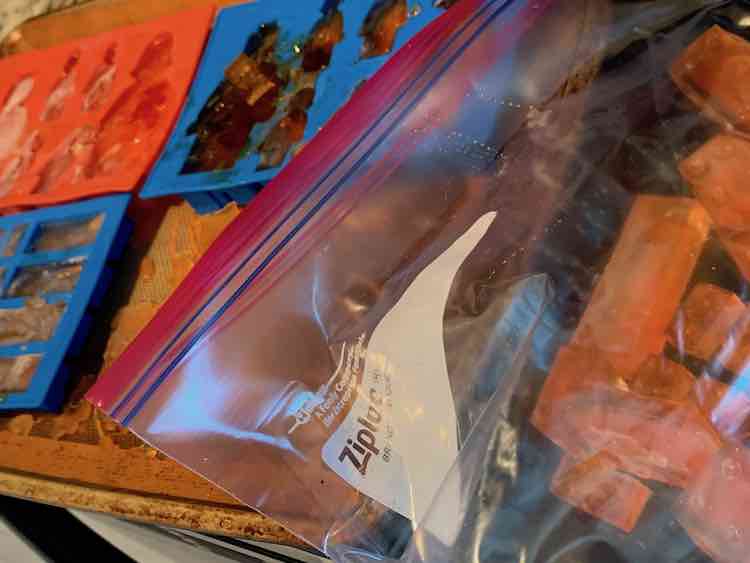 Foreground Ziploc freezer bag with a cookie sheet containing the other three silicone moulds filled with orange dyed ice in the background. 