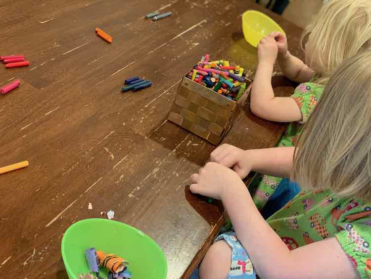 Both kids at the table with a container of crayons between them and each has a bowl for the paper scraps. They've separated the peeled crayons based on their hue.