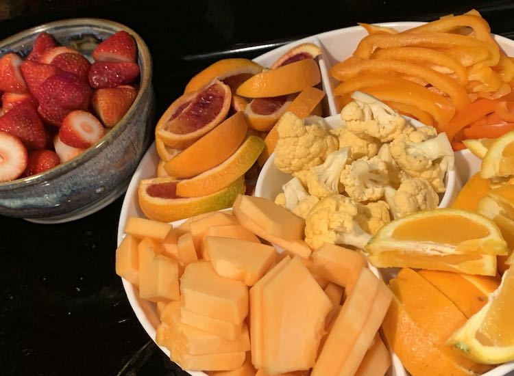 Mostly orange themed fruits and veggies. Tray holds the orange and there's a bowl of red strawberries in the back.