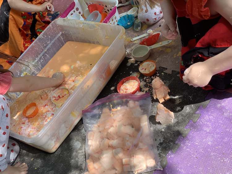 Kids gathered around a bin of orange oobleck with flecks or red mixing in. Bag of frozen oobleck in the foreground.