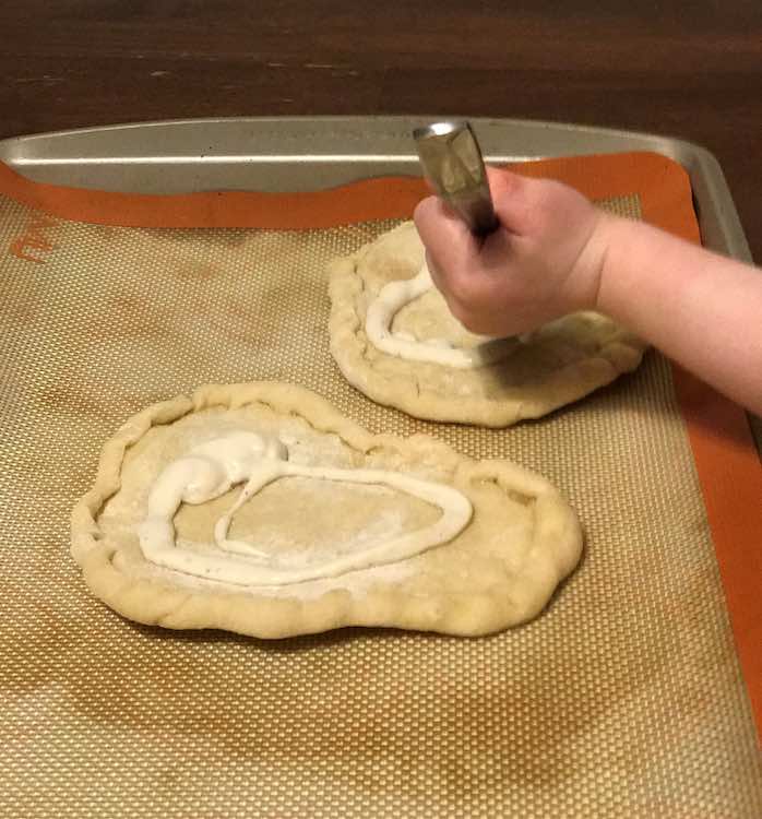 Closeup of Ada spreading ranch dressing on her pizza crust with a spoon. Underneath the two pizza crusts are a silpat and cookie sheet. 