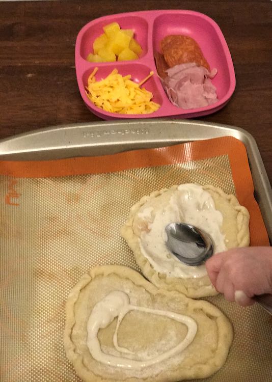 While Ada still spreads the ranch dressing on the pizzas on the cookie sheet there's a plate of toppings in the background with grated cheese, pineapple, pepperoni, and ham sandwich meat. 