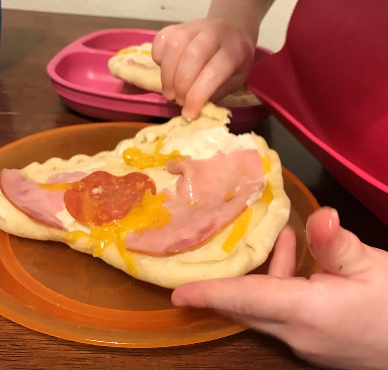 The one pizza sits on an orange plate and the edge is slightly lifted up by two toddler hands. The pizza between the two hands has a little nibble already stolen off. In the background the other pizza sits on the plate that used to house the toppings.