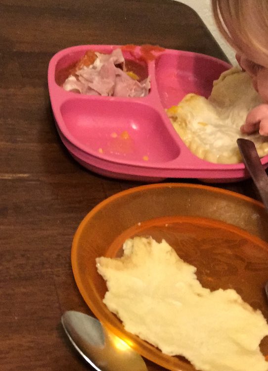 The orange plate in the foreground is now naked with bites taken around the outside. Behind the plate is another, pink sectioned, plate with the meat toppings filling one of the smaller sections. Ada leans over to take a bite of the other crust.