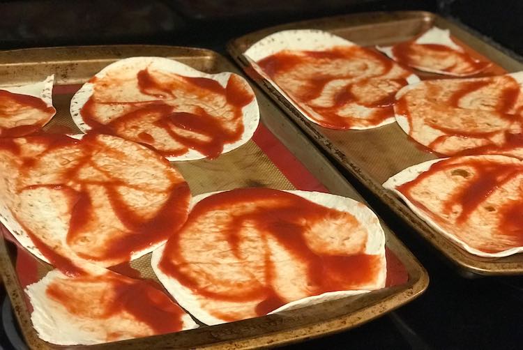 Closeup of the two cookie sheets with the tomato sauce covered flour tortillas. 