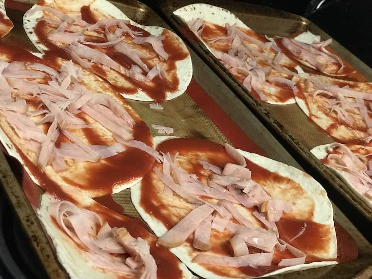 Closeup view of the cookie sheets, angled from the side, showing them with tomato sauce and strips of deli meat on top.