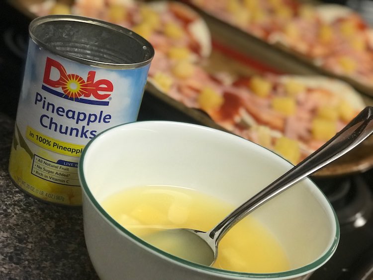 Closeup of a white bowl with a spoon, inside it, sitting in yellow liquid with the odd floating chunk of pineapple. Behind the bowl is the opened and emptied can of pineapple chunks and behind both are the blurred out pizzas from before now also topped with the pineapple chunks.