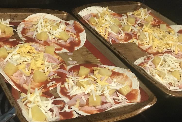 View angled from above of the two cookie sheets with tortilla wraps topped with tomato sauce, deli meat, pineapple chunks, and mixed grated cheese. 