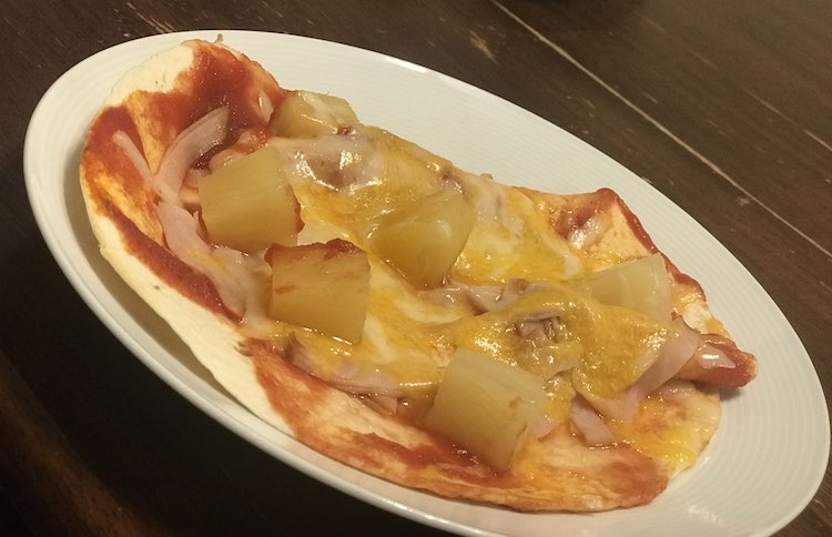 Image of a personal pizza topped with melted cheese and other toppings sitting on a white plate on a wooden table. 