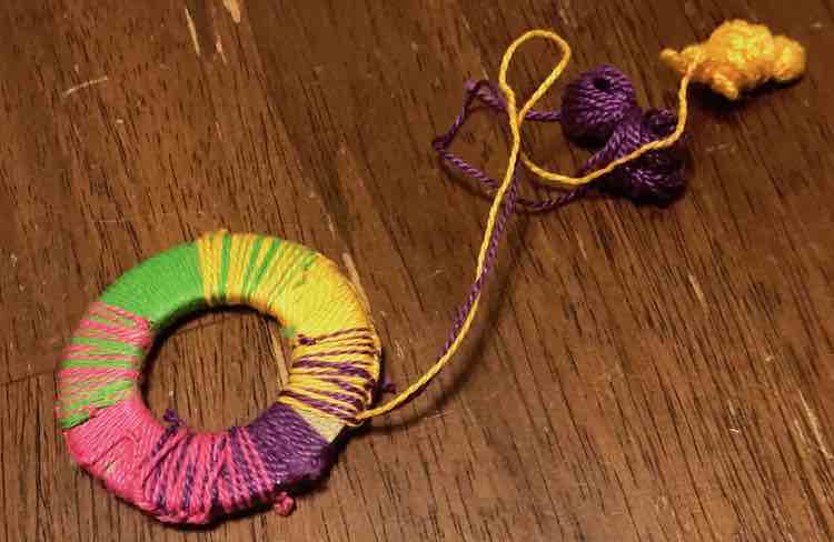 The friendship bracelet washer is almost done with both yellow and purple embroidery floss leading away from the minor empty space left on the washer. 