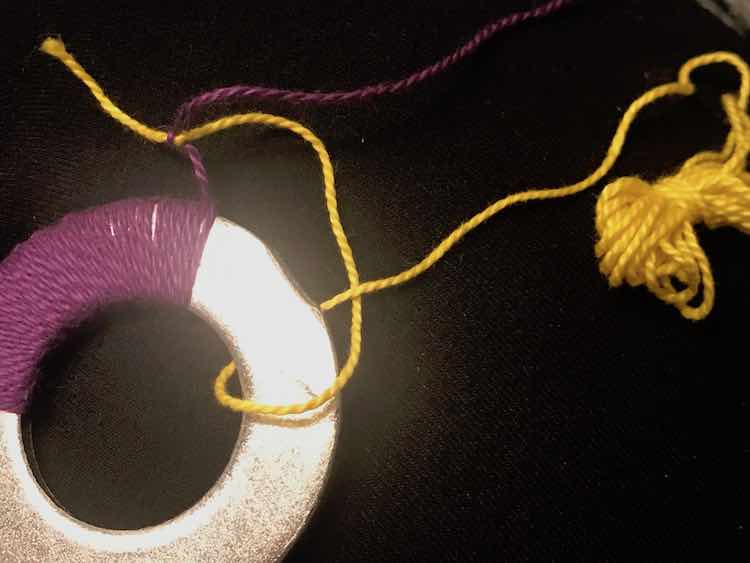 Washer with purple embroidery floss wrapped around part of it. The end of the purple floss is tied to a new ball (yellow) which is then loosely wrapped around the washer and pulled through the loop making a loose knot.