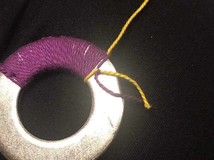 Washer with purple embroidery floss knotted on it and a single strand of yellow knotted. The purple/yellow knot is showing on the last loop around and the loose ends dangle over the naked washer.