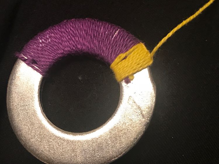 Large swash of purple embroidery floss looped around the washer next to a small section of yellow. At the edge of the yellow section you can see the purple and yellow tips of the loose ends sticking out. The next loop/knot around the washer will hide them.