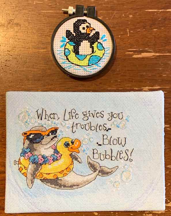 The two finished and mounted embroidery kits. The smaller circled one (penguin floating in an inner tub) above the dolphin in an inner tube with quote: "When Life give you troubles... ... Blow bubbles!"
