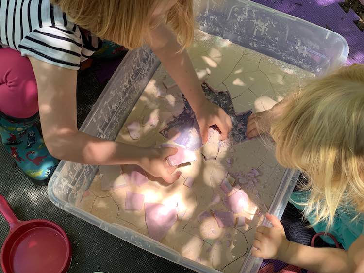 Closeup of a white bin filled with cracked oobleck. The kids are leaned over either side picking up flat pieces of oobleck showing the pink underside. The top is a yellow orange depending on where you look.