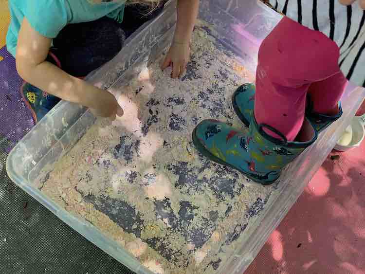 Zoey crouches by the bin of dried out sand as Ada stand within it. Two different methods for two different kids.