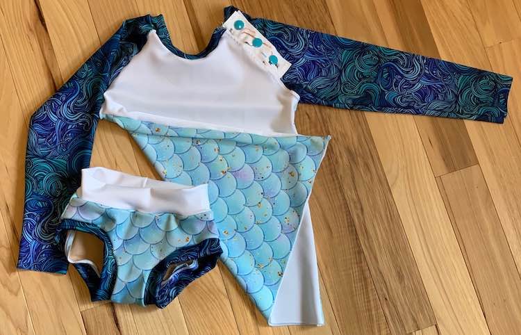The front of the swim bottoms are showing while the bottom of the swimming top is folded over hiding the mermaid but showing the scales on the back of the suit. 