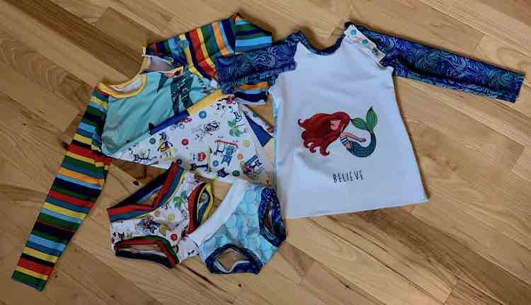 Image shows a flat lay of two swimming suits. Both suits consist of a raglan and a pair of bottoms. The one is Pete the cat with stripes and a panel on the front. The other is a mermaid saying "believe" and coordinating scales and waves. 