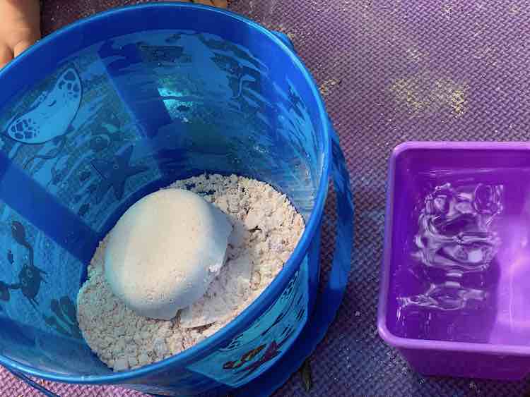 Blue pail with oobleck sand in it and a purple container of water.