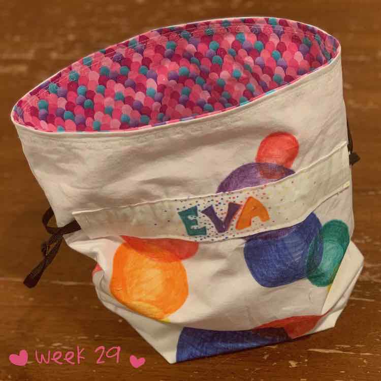 Flat bottom drawstring bag. The inside is made from sparkly scale fabric. The outside is white with colored spheres drawn on. The drawstring is kept in place with some fabric showing the recipient's name.