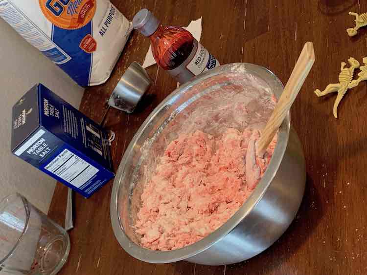Large metal bowl of mostly mixed red-colored salt dough with the red dye, flour, salt, measuring cups, and dinosaur fossils in the background.