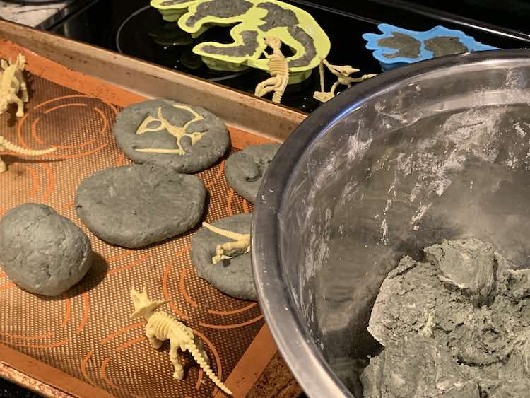 Bowl of unused grey salt dough in the foreground with fossil imprinted rocks forming themselves in the background. Behind all of that you can see the dinosaur skeleton moulds filled with grey salt dough.