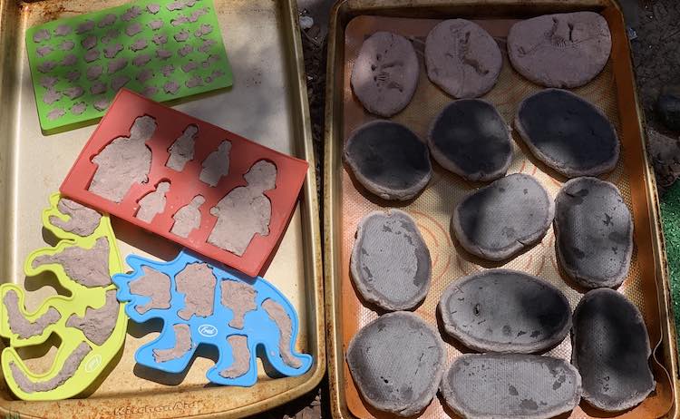 One cookie sheet, Silpat, and fossil imprinted rocks on the right (most of them flipped upside down showing their wetter side) and the silicone moulds filled with grey salt dough and corralled on another cookie sheet to the left.