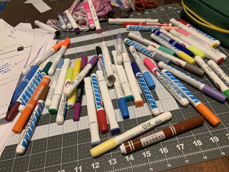The fabric markers that match are set out of the way in the background while the rest of the markers are spread out on a cutting mat in the foreground. The pencil bag that normally holds them are off to the side to the right. 