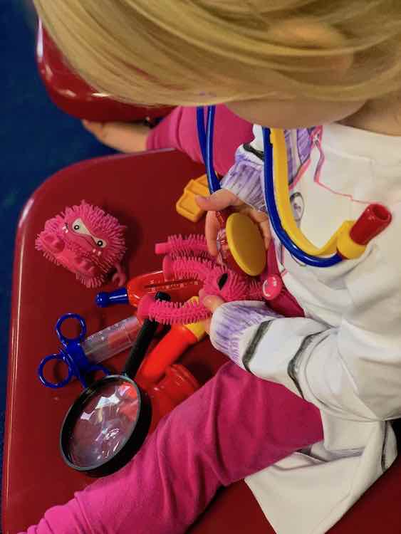 Zoey sitting on a red chair with two squeeze toys (one missing a leg) and her doctor tools. She's giving a checkup to the monster missing a leg.
