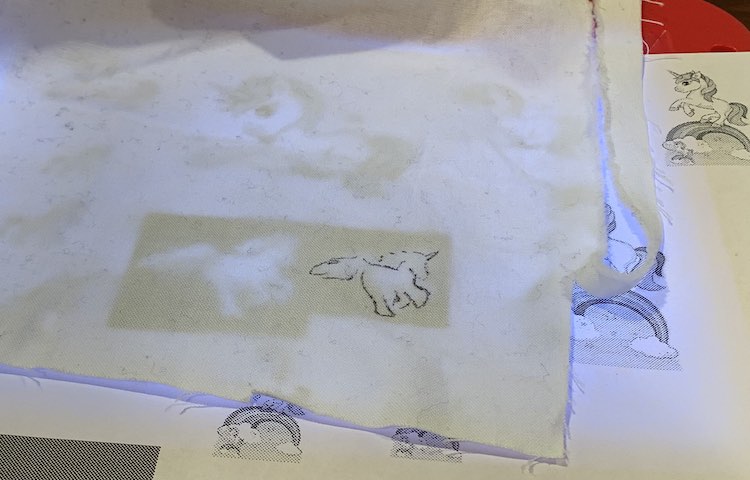 White woven fabric with a running unicorn partly traced. The fabric is draped over a piece of printed paper and a kids’ light box.