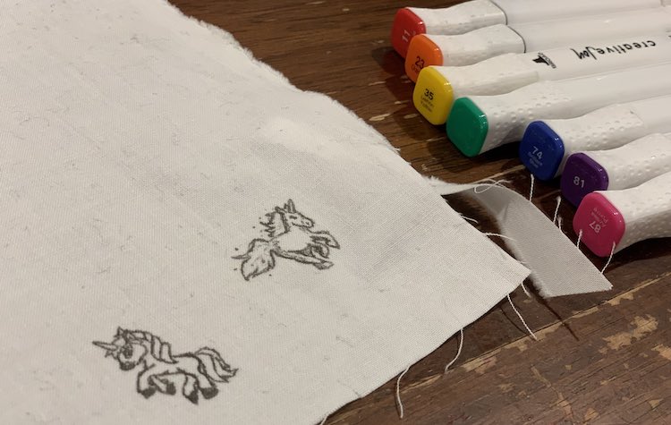 Corner of white woven fabric showing two white unicorns outlined and detailed with a fine-tipped black fabric marker. An array of rainbow colored fabric markers are lined up next to it ready to go. 