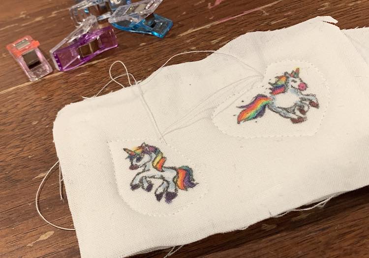 A swatch of fabric with two unicorns colored on. The left unicorn has a tear drop sewn around it while the right most unicorn has a heart. Both shapes have the threads left untrimmed at this point. 