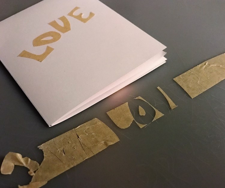 The masking tape is shown on the cutting board with the letters from "LOVE" removed yet the WE showing on the left and LOVE disappearing off the right side. The white card is shown above the tape and the word LOVE has already been moved up.