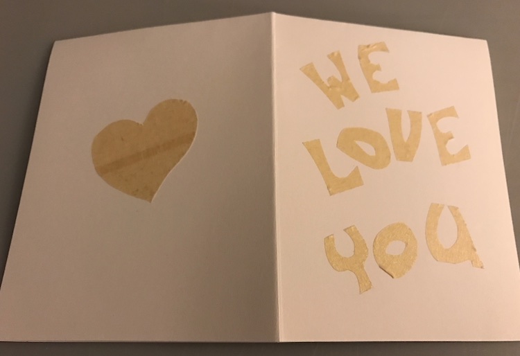 Card is opened and inside down so you can see the back (left side) and front (right side) of the card. The back has a heart and the front says "we love you" both in masking tape stuck to the paper.