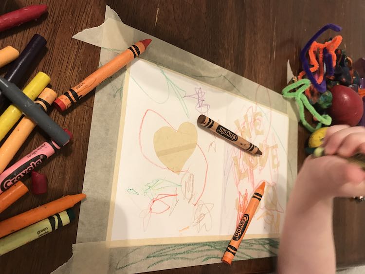 Ada with a green crayon poised about to color on the taped on card on the table. The card is taped to the table underneath her hand with a pile of crayons to the left. Pipe cleaners, inexplicably, to the right along with egg shaped crayons. 