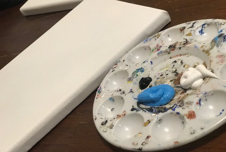 Right foreground holds a used and dried plastic paint palette with three dollops of paint. The larges dollop is blue, then white, and finally a small dab of black. To the left in the background sits a canvas.